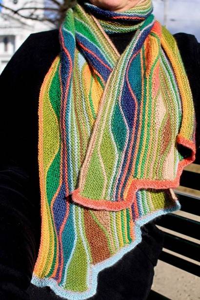 a woman is shown wearing black top with a colorful rectangular stole/scarf wrapped around her neck. The scarf has waving blocks of colors including blue, teal, green, yellow, and coral. Each block is separated by a narrow line of contrasting color. The whole piece is knitted in garter stitch